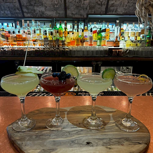 Four colorful cocktails on a wooden board at a bar, with a well stocked liquor shelf in the background, lime garnish on each.