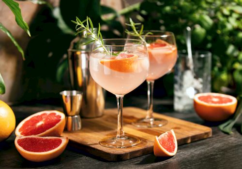 Two elegant grapefruit cocktails on a wooden board, garnished with herbs, sliced grapefruit nearby in a sunny garden setting.