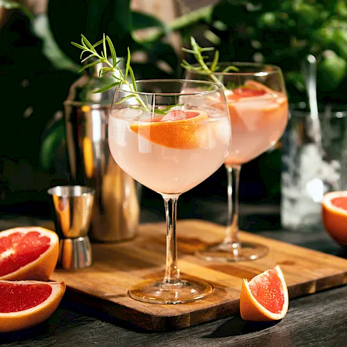 Two elegant grapefruit cocktails on a wooden board, garnished with herbs, sliced grapefruit nearby in a sunny garden setting.