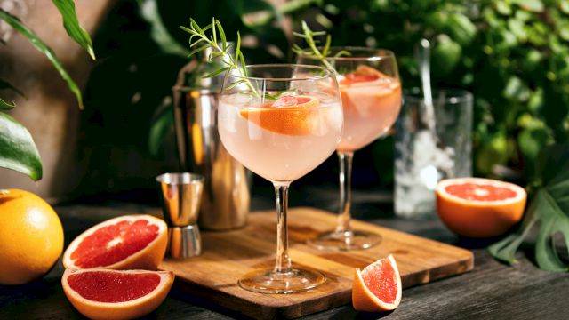 Two elegant grapefruit cocktails on a wooden board, garnished with herbs, sliced grapefruit nearby in a sunny garden setting.