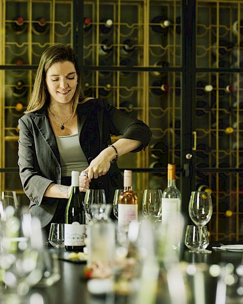 A woman leads a wine tasting at a table with bottles, glasses, and a wine rack backdrop, guiding the group through the event.