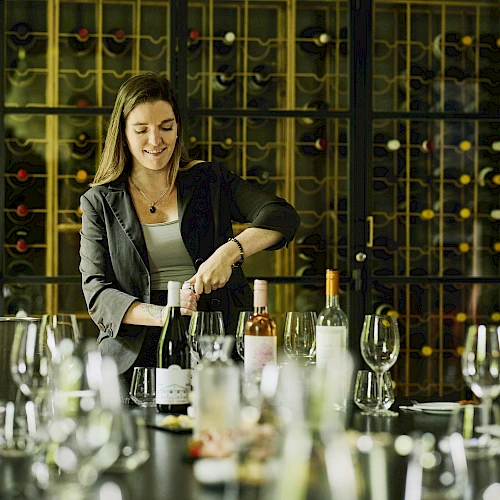 A woman leads a wine tasting at a table with bottles, glasses, and a wine rack backdrop, guiding the group through the event.