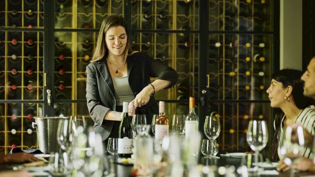 A woman leads a wine tasting at a table with bottles, glasses, and a wine rack backdrop, guiding the group through the event.