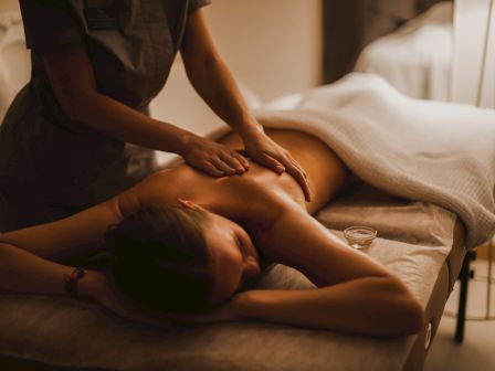 A person receives a back massage in a dim spa room, lying face down while a therapist works on their shoulders and back, relaxing ambiance.