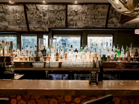 A well-lit bar with a long counter full of assorted liquor bottles, glassware, and stools, illuminated by warm overhead lights at night.