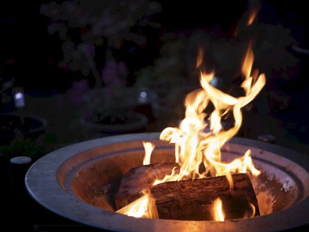 A glowing fire pit with burning logs crackles in a round metal bowl against a dark background.