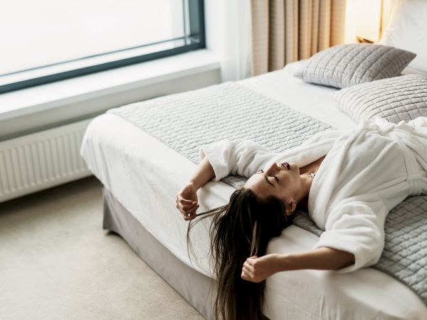 A woman lies on a neatly made bed in a bright, minimalist bedroom with a large window and soft cushions, relaxing in a white robe.
