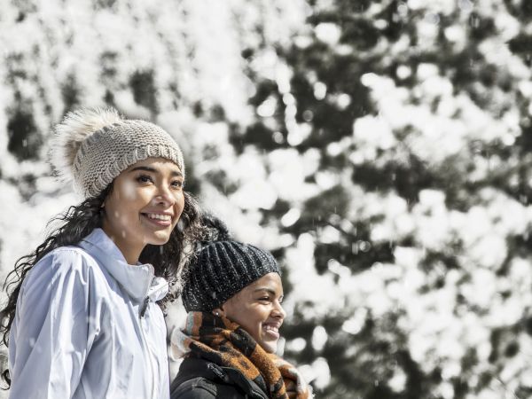 Two people smile in winter coats and knit hats, standing outdoors with snowy trees in the background, enjoying a chilly day together.