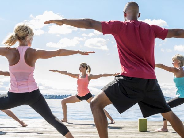 A group of people doing a lakefront yoga or martial-arts-style pose on a wooden pier, arms spread wide, in bright workout attire.