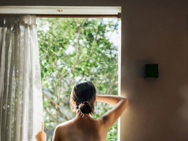 Woman standing at an open door, back to camera, looking outside at greenery. Curtains drift in, sunlight spills in, casual hair bun.