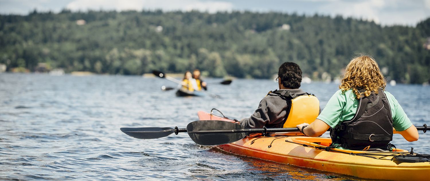 Two people wearing life jackets kayak on a calm lake, with others paddling in the distance under a cloudy sky.