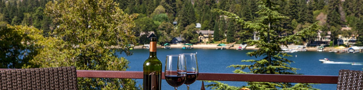 A lakeside table setup with wine, glasses, and a cheese platter on a wicker patio, overlooking a forested shoreline and blue water.