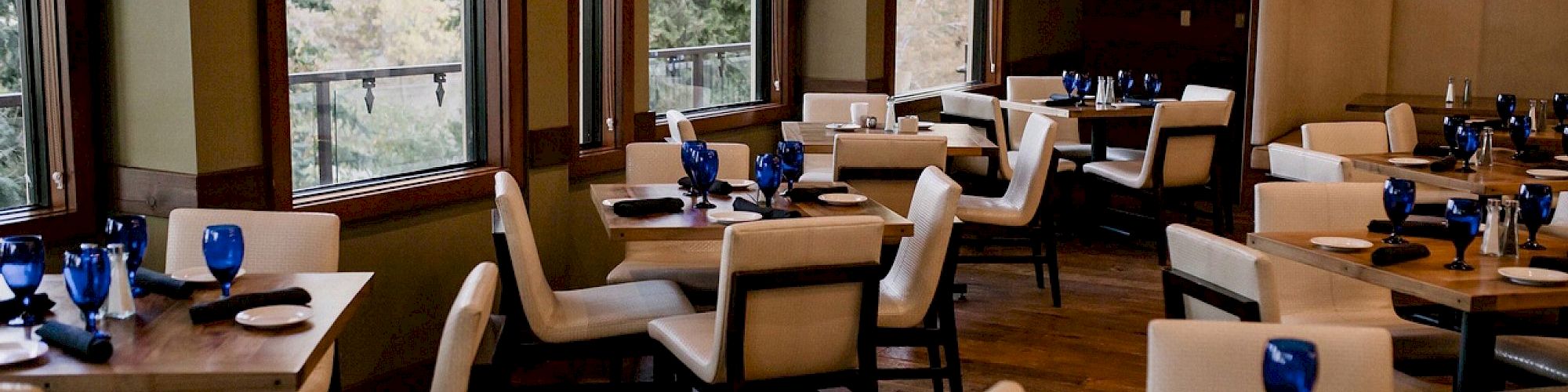 A cozy restaurant with wooden floors, large windows, cream chairs, and tables set with blue glassware and white plates, ready for guests.