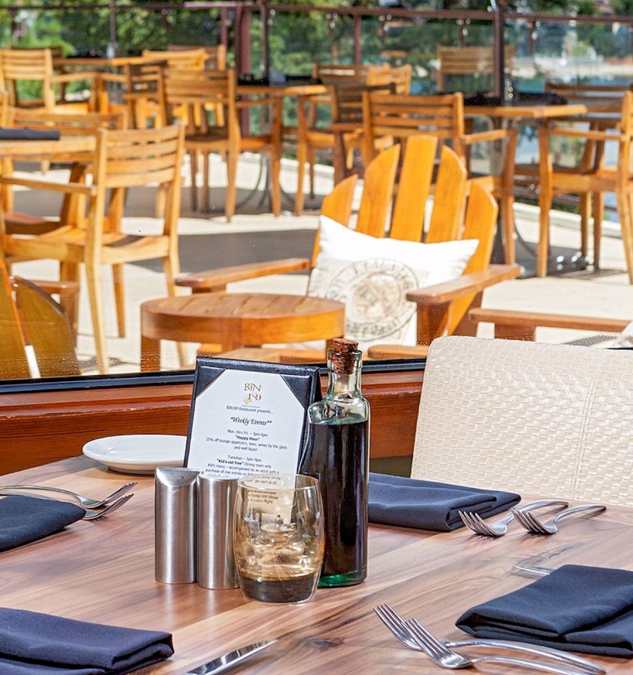 A cozy outdoor restaurant with wooden tables and chairs, navy napkins, and a bottle of oil or sauce on the table, sunny patio vibes.