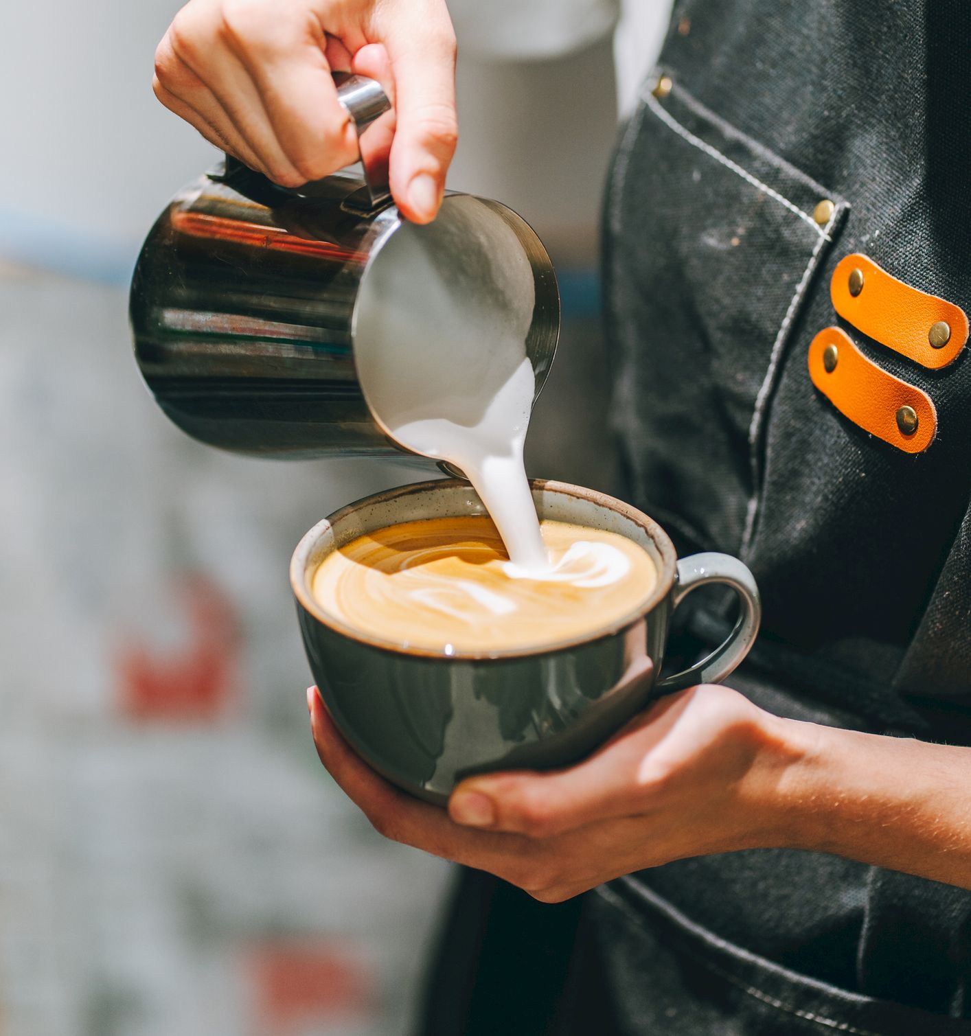 A barista pours steamed milk into a cup of coffee, creating latte art on top.