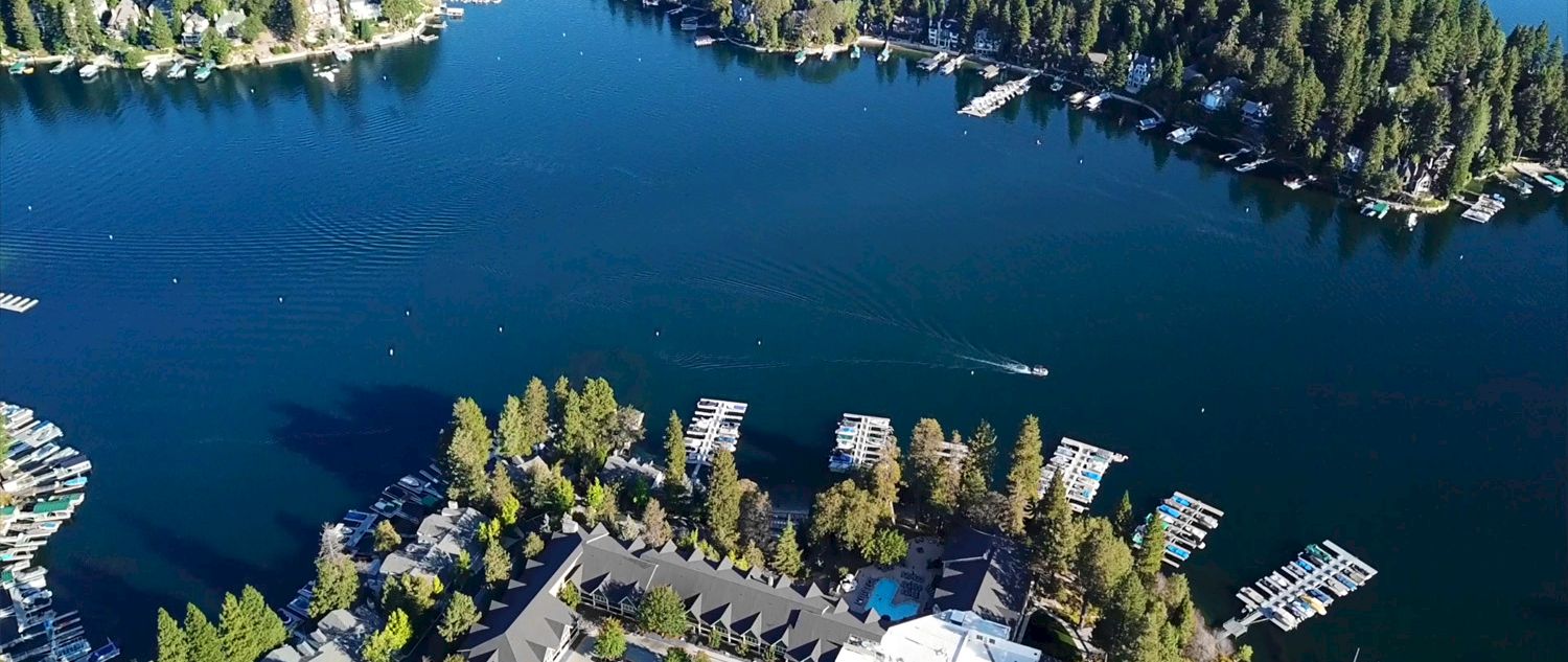 Aerial view of a forested lakeside resort with docks, boats, and cabins along a calm blue lake.