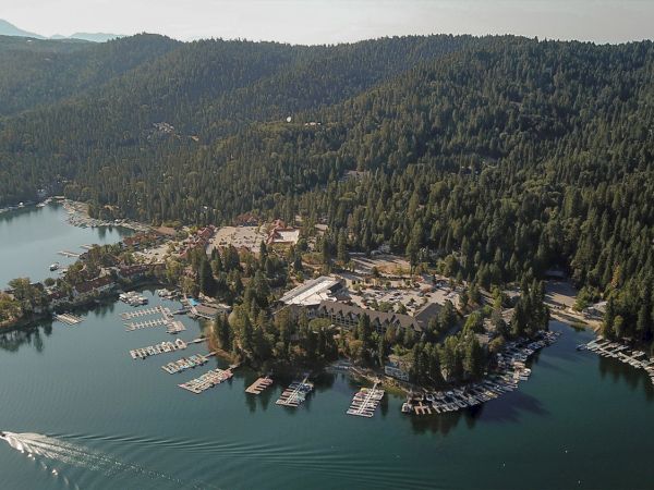 Aerial view of a lakeside village with marinas and many docks, surrounded by dense green forested hills and mountains.