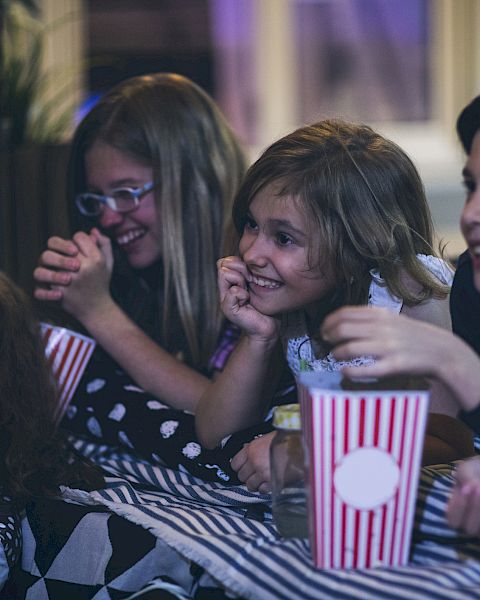 A group of friends at a party huddle together, laughing and eating popcorn from striped cups, looking excited and happy.