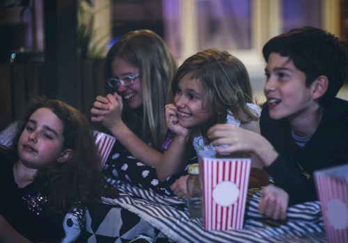 A group of friends at a party huddle together, laughing and eating popcorn from striped cups, looking excited and happy.