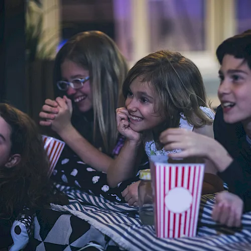 A group of friends at a party huddle together, laughing and eating popcorn from striped cups, looking excited and happy.