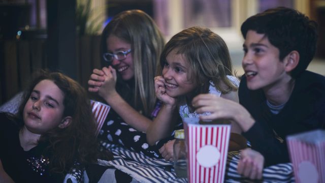 A group of friends at a party huddle together, laughing and eating popcorn from striped cups, looking excited and happy.