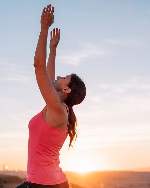 A woman in a pink top is stretching her arms overhead outdoors at sunrise, reaching toward the sky in a calm moment of exercise.