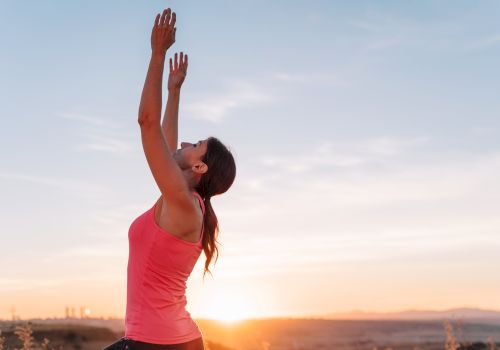 A woman in a pink top is stretching her arms overhead outdoors at sunrise, reaching toward the sky in a calm moment of exercise.