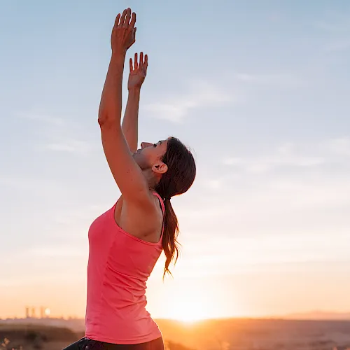 A woman in a pink top is stretching her arms overhead outdoors at sunrise, reaching toward the sky in a calm moment of exercise.