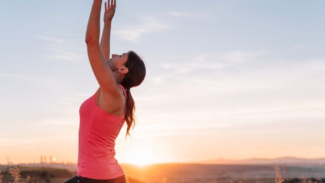 A woman in a pink top is stretching her arms overhead outdoors at sunrise, reaching toward the sky in a calm moment of exercise.