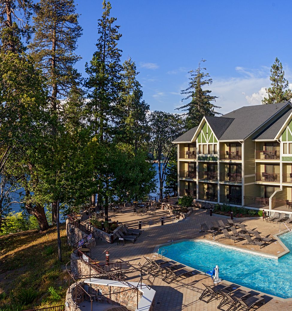 A lakeside resort with a large pool, lounge chairs, and a multi-story building surrounded by trees under a blue sky.