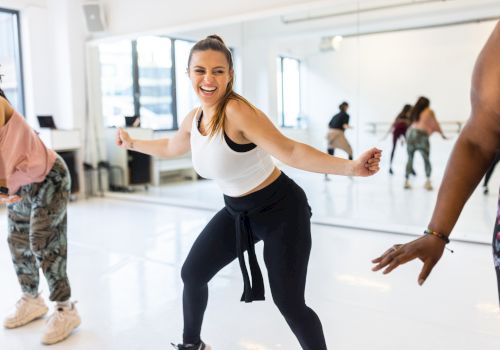 In a bright fitness studio, a woman leads a joyful workout dance while others stretch and move nearby.