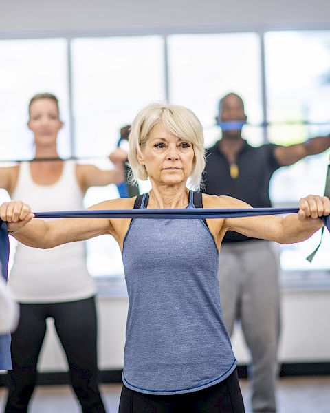 An older woman leads an exercise class, holding a resistance band while others stretch in a bright gym room.