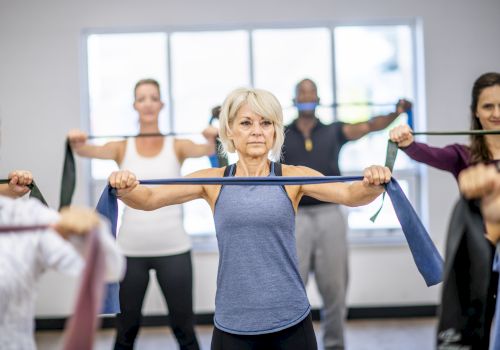 An older woman leads an exercise class, holding a resistance band while others stretch in a bright gym room.