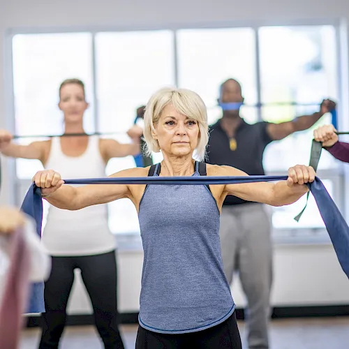 An older woman leads an exercise class, holding a resistance band while others stretch in a bright gym room.