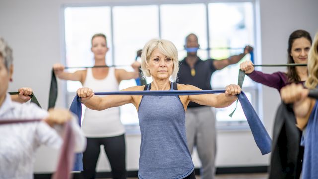 An older woman leads an exercise class, holding a resistance band while others stretch in a bright gym room.