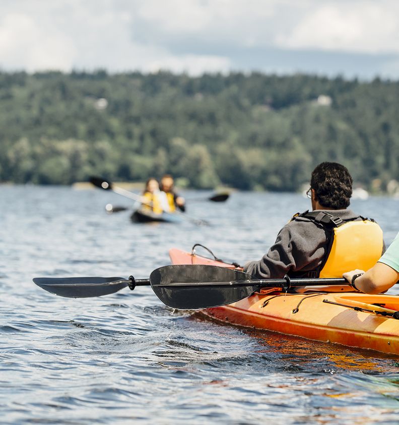 Two people paddle a bright yellow kayak on a lake, with more kayakers ahead and a forested shore in the background.
