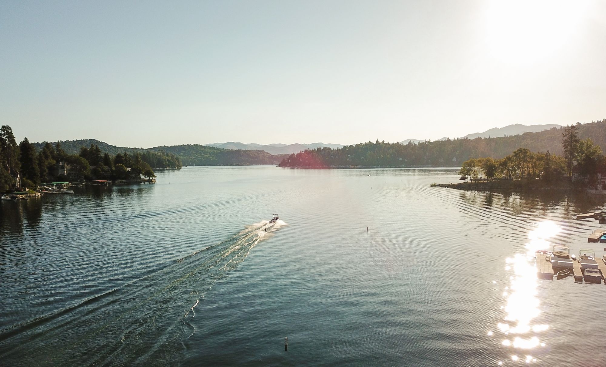 A wide calm lake at sunset with trees and houses along the shores, and a speedboat cutting ripples across the water.