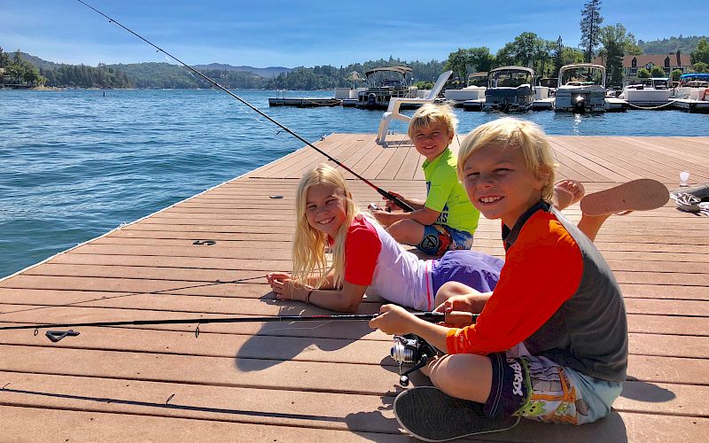 Three kids smile on a sunny boat dock by a lake, with a fishing rod extended over calm water.