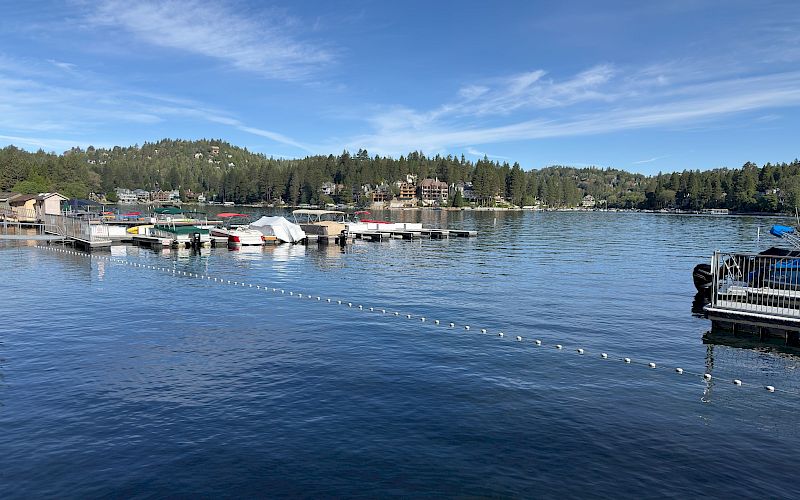 A peaceful lakeside marina with boats moored at docks, calm water, and a tree-lined hillside under a blue sky.