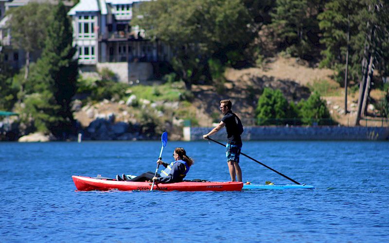 Two paddlers glide in a red kayak on blue water with a green forest and a building on the shore behind them.