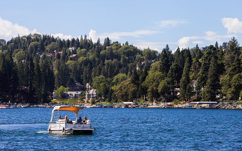 A sightseeing boat with people glides on a blue lake by a marina, with green wooded hills under a sunny sky.