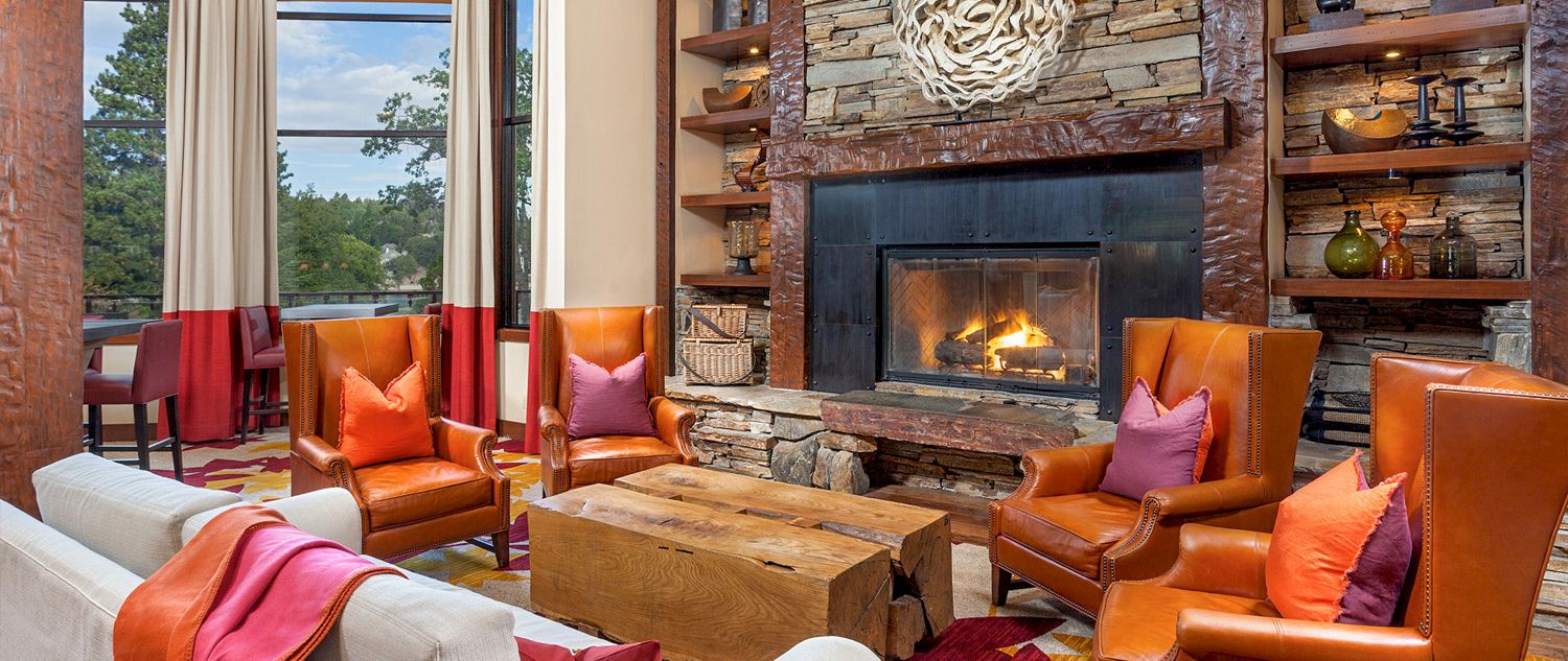 A cozy living room with a stone fireplace, leather armchairs, a rustic wooden coffee table, and warm decor by big windows.