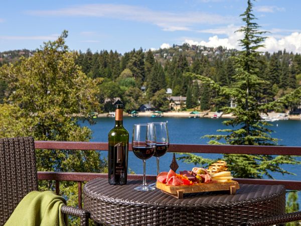 A lakeside patio table shows a wine bottle, two glasses, and a cheese platter with the lake and trees behind it.