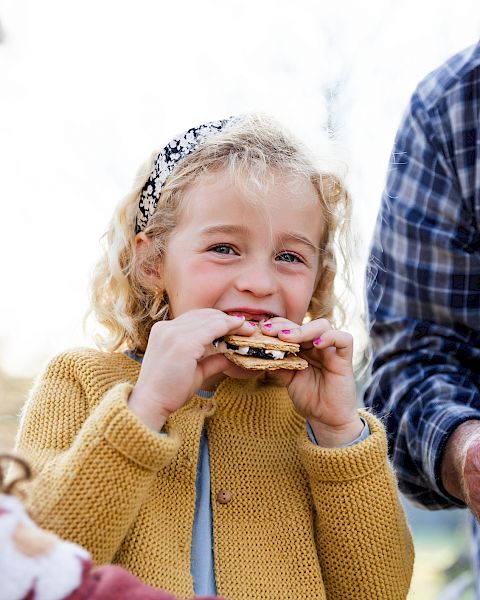 A little girl happily eats a cracker/cookie outdoors with older family members nearby, all smiling and sharing the moment.