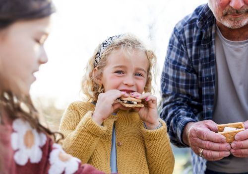 A little girl happily eats a cracker/cookie outdoors with older family members nearby, all smiling and sharing the moment.