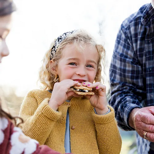 A little girl happily eats a cracker/cookie outdoors with older family members nearby, all smiling and sharing the moment.