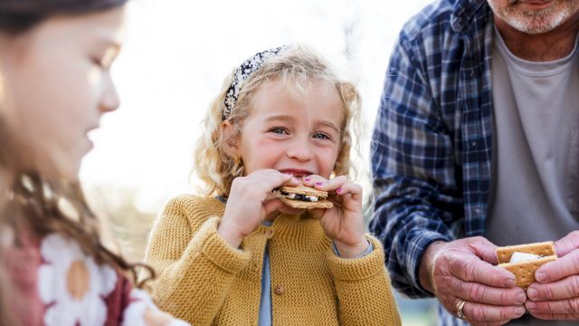 A little girl happily eats a cracker/cookie outdoors with older family members nearby, all smiling and sharing the moment.
