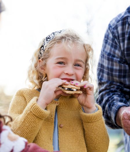 A little girl smiles while holding a cracker sandwich as an older couple shares treats outdoors.