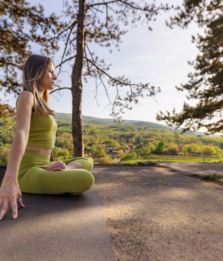 A woman in yellow yoga pants meditates outdoors on a stone ledge, surrounded by trees and hills at golden hour.