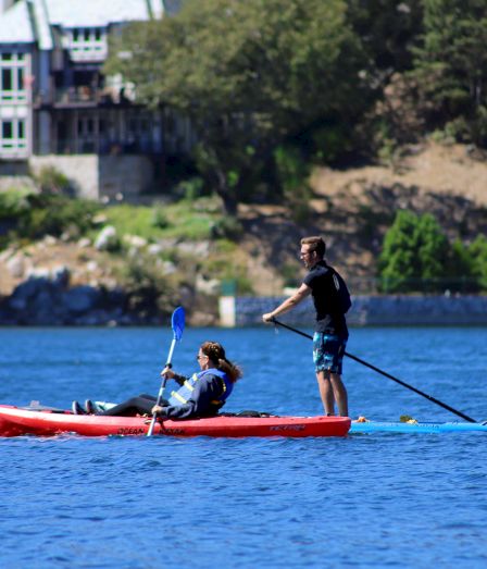 Two people paddle a red ocean kayak on a blue lake while a house and trees line the shore behind them.
